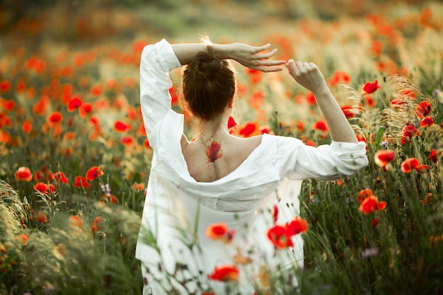 Fille avec un dos nu avec un tatouage dessus se tient la main sur la tête et le magnifique champ de coquelicots