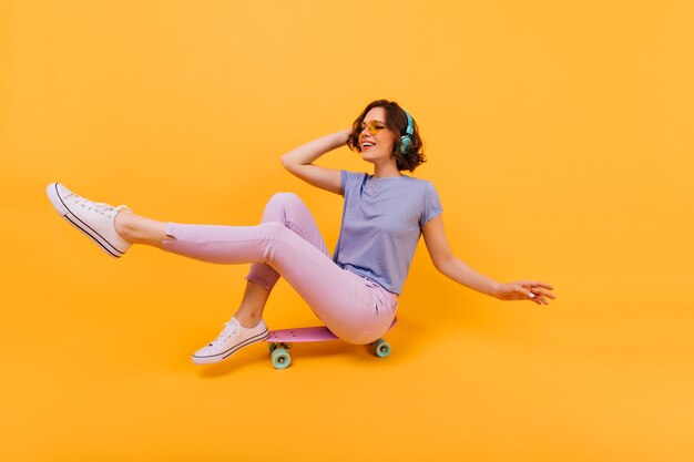 Fille drôle en pantalon rose assis sur une planche à roulettes et faire des grimaces. Photo intérieure de la belle femme blanche dans les écouteurs