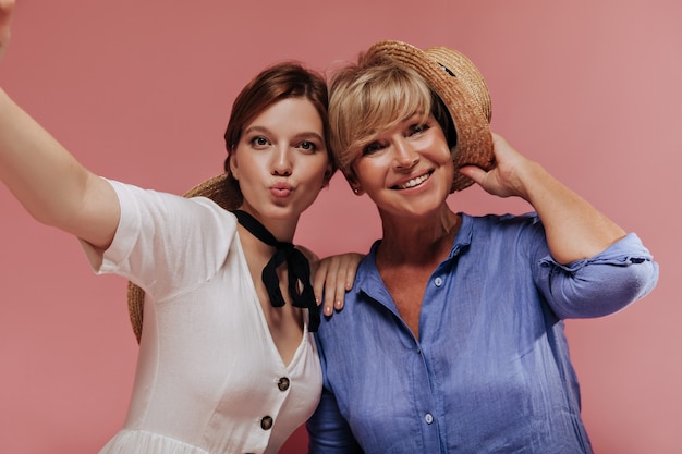 Fille drôle en robe blanche faisant selfie avec une femme heureuse aux cheveux courts blonds en tenue bleue et chapeau de paille sur fond rose.