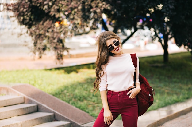 Fille efficace avec de longs cheveux bouclés en pantalon vineux pose dans la rue de la ville.