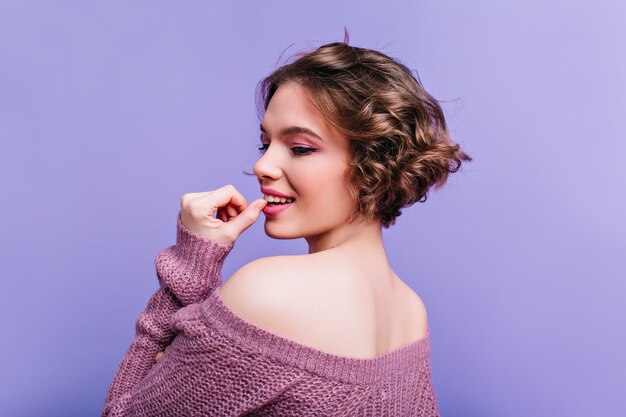 Fille effrayée en écharpe tricotée blanche couvrant le visage avec les mains. Portrait intérieur d'une jeune femme européenne émotionnelle au chapeau isolé sur un mur violet.