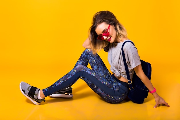 Fille élégante dans des verres lumineux avec sac à dos assis sur le sol avec les yeux fermés. Portrait en studio de jeune femme en chaussures de sport et leggings isolés sur fond jaune.