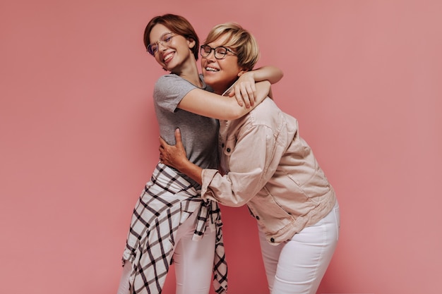 Fille élégante dans des vêtements à carreaux légers souriant et serrant la vieille femme dans des verres une veste beige fraîche sur fond rose isolé.