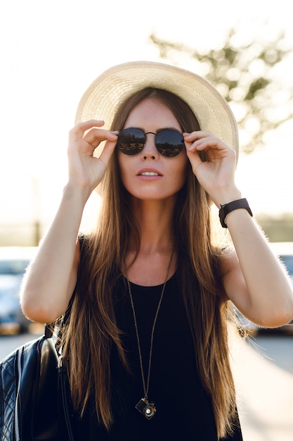 Photo gratuite fille élégante debout près de la route portant une robe noire courte, un chapeau de paille, des lunettes noires et un sac à dos noir. elle sourit sous les chauds rayons du soleil couchant. elle touche ses lunettes de soleil avec la main