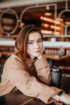 Une fille élégante est assise dans un café à table et boit du café. café à emporter dans un gobelet en carton. femme aux cheveux roux dans un costume chaud beige dans une atmosphère chaleureuse. intérieur moderne. passe-temps calme et agréable.