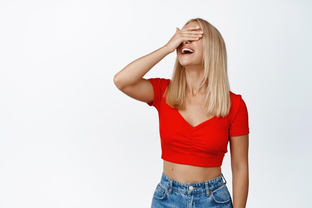 Fille élégante insouciante aux cheveux blonds couvre ses yeux les yeux bandés rit et sourit attend des stands de surprise sur fond blanc