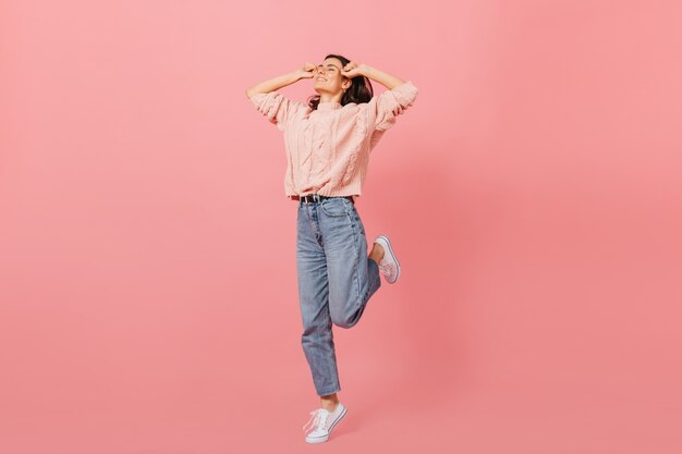 Fille élégante en jeans de maman et pull surdimensionné chaud sautant joyeusement sur fond rose.