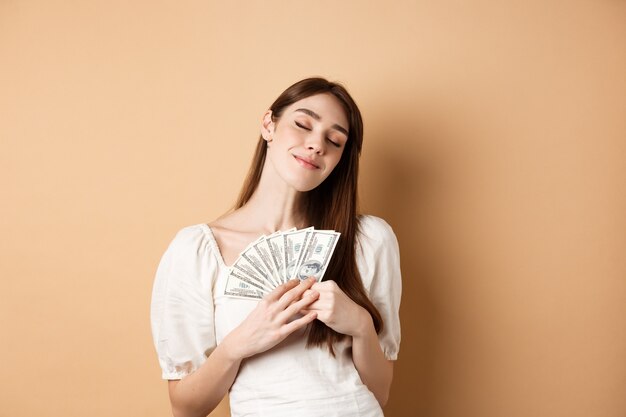 Fille élégante serrant de l'argent avec un sourire heureux et des yeux fermés comme des billets d'un dollar debout sur un ba beige ...