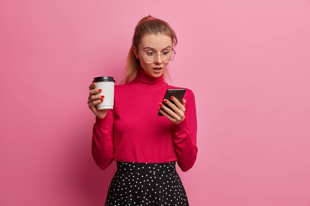 Une fille émotionnelle choquée regarde l'écran du smartphone, discute avec des amis en ligne, porte de grandes lunettes optiques, tient une tasse jetable de boisson fraîche, apprécie un café savoureux