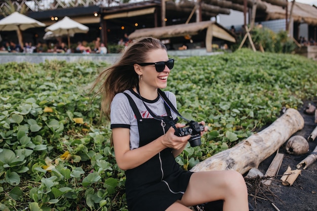 Fille émotionnelle en tenue vintage assis sur la pelouse avec appareil photo. Rire femme photographe en lunettes de soleil noires s'amuser dans le parc en journée d'été.