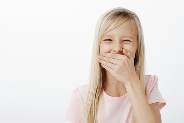 Photo gratuite fille émotive a appris un mot drôle. joyeux adorable enfant européen aux cheveux blonds, couvrant la bouche de paume et riant aux éclats, entendant une blague hilarante ou regardant un dessin animé