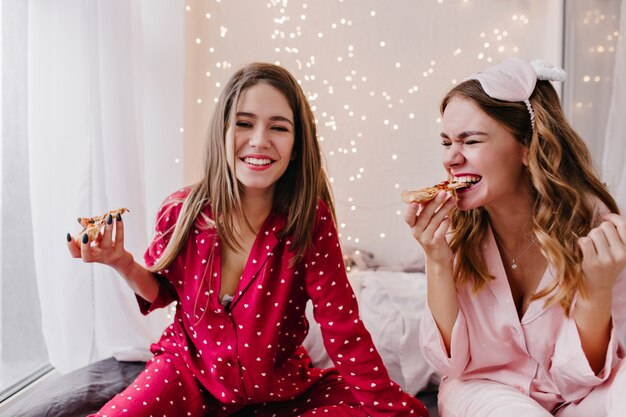 Fille enthousiaste avec une coiffure frisée, manger de la pizza. Photo intérieure d'une jeune femme caucasienne heureuse en pyjama rouge posant dans la chambre avec restauration rapide.