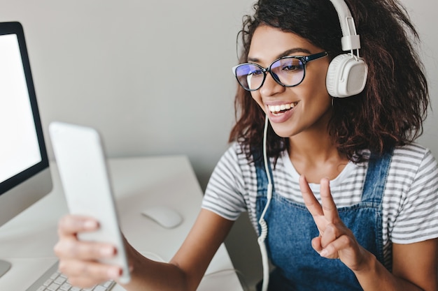 Photo gratuite fille enthousiaste portant des lunettes faisant selfie avec signe de paix passer du temps au bureau