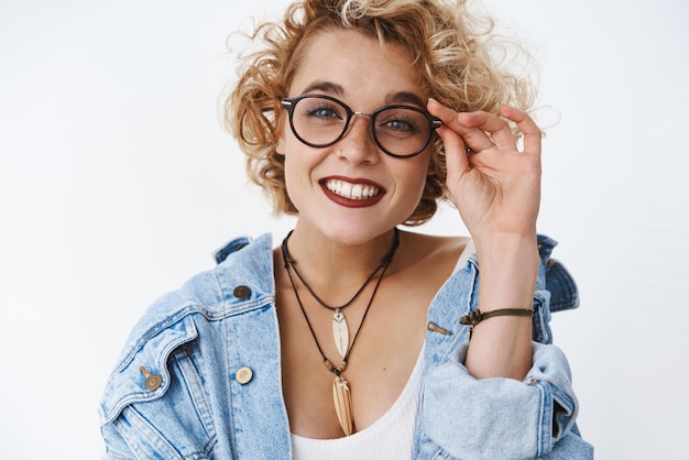 Une fille enthousiaste à ce rendez-vous aime vraiment une conversation intéressante touchant le bord des lunettes et souriant largement avec une expression curieuse et amusée écoutant une histoire géniale debout heureux sur un mur blanc