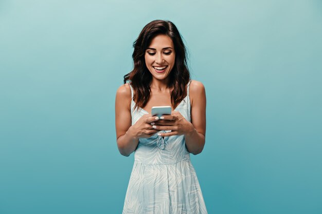 Fille espiègle en robe blanche avec sourire bavardant dans un smartphone sur fond bleu. Enthousiaste belle femme de bonne humeur tient le téléphone.