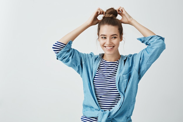 La fille essaie une nouvelle coiffure. Projectile studio, de, joyeux, émotif, européen, girl, confection, chignon, sur, cheveux, et, regarder côté, miroir, sourire positivement, quoique, debout, dans, élégant, équipement