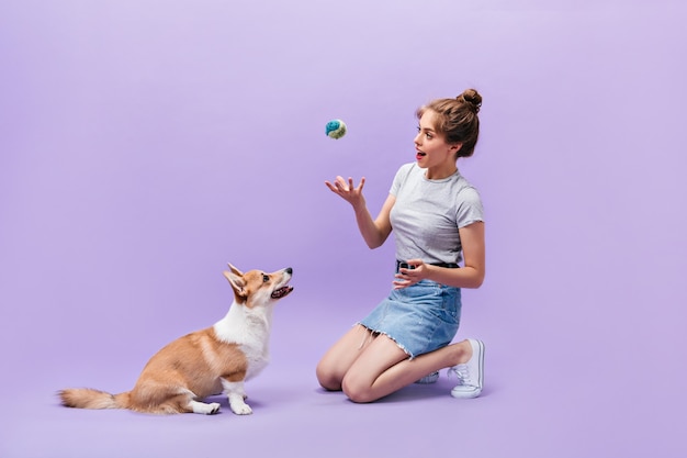 Photo gratuite fille est assise sur le sol et joue avec le chien. heureuse jeune femme en baskets blanches posant avec ballon et corgi sur fond violet.