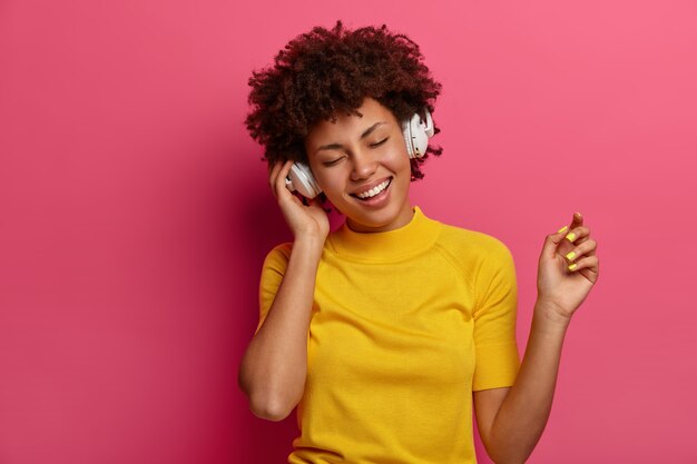 Une fille ethnique optimiste bouge sans soucis et écoute de la musique dans un casque, se sent détendue, apprécie la mélodie préférée ou une nouvelle piste sur l'application de la chanson, porte des vêtements jaunes, isolés sur un mur rose. Technologie, gadgets