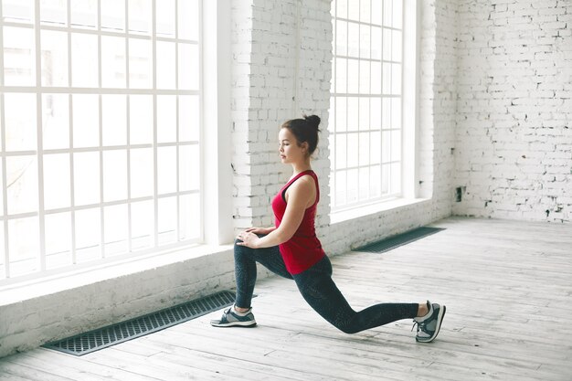 Fille étudiante déterminée et confiante avec noeud de cheveux faisant de l'exercice physique à l'intérieur avant l'université. Élégante jeune femme sportive en baskets et vêtements de sport debout en fente basse, étirement des muscles des jambes