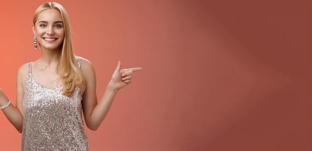 Une fille européenne blonde et joyeuse en robe argentée étincelante pointant sur le côté.