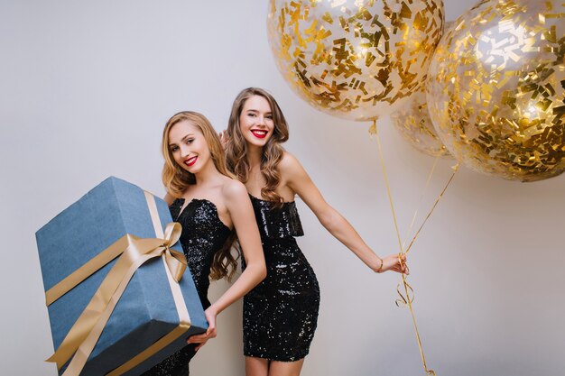 Fille européenne bouclée à la mode danse avec des ballons derrière une amie blonde. Photo intérieure de joyeuse jeune femme tenant un cadeau décoré de ruban et souriant doucement.