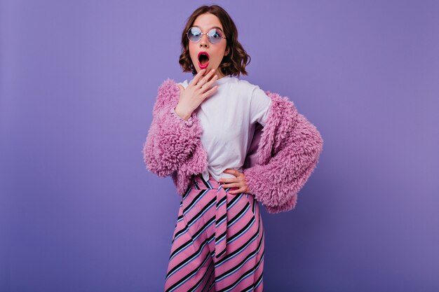 Fille européenne choquée en t-shirt blanc et manteau de fourrure rose posant. Photo intérieure d'une jolie femme avec une coiffure courte exprimant son étonnement sur un mur violet.