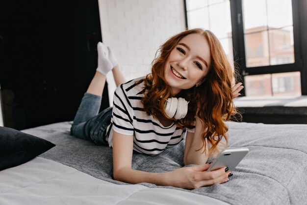 Fille européenne glamour à l'aide de téléphone tout en posant sur le lit. Femme au gingembre de bonne humeur dans les écouteurs s'amusant le week-end.