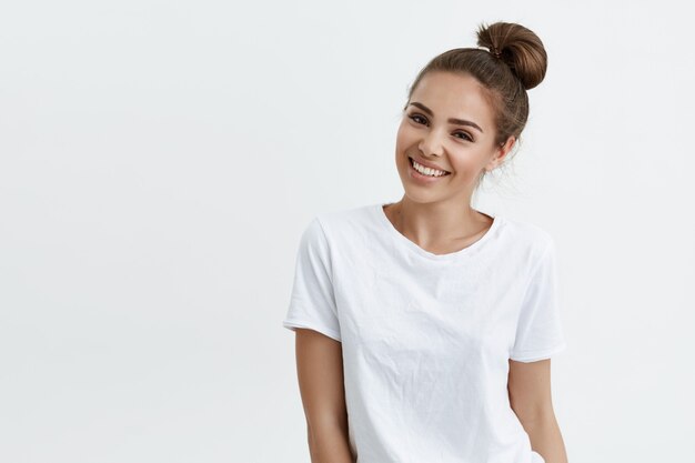 Fille européenne mince positive avec une coiffure en chignon, souriant largement en se tenant debout sur un espace blanc, exprimant la confiance et la sensualité.