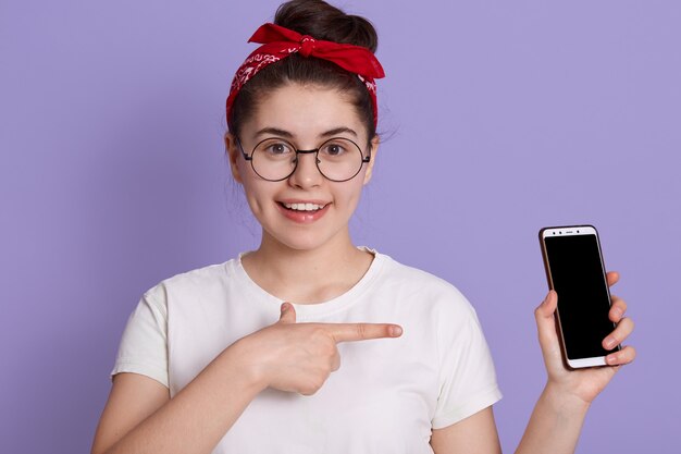 Fille européenne montrant l'écran du téléphone portable vierge avec le doigt avant et avec un sourire charmant, femme avec un t-shirt décontracté blanc et un bandeau rouge