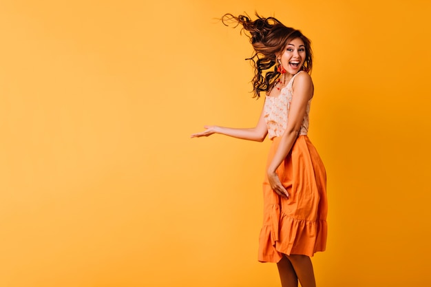 Fille excitée aux cheveux ondulés au gingembre sautant sur le jaune. Portrait en studio d'une jeune femme heureuse en tenue orange dansant avec le sourire.