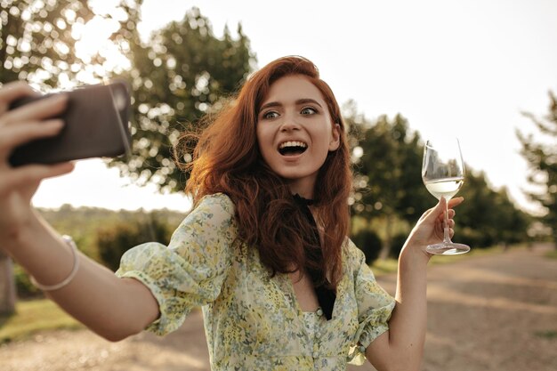 Fille excitée avec des cheveux roux et un bandage noir sur le cou en robe jaune tenant un verre avec du vin et prenant un selfie en plein air