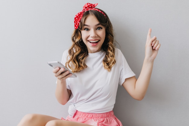 Fille excitée avec joli ruban rouge posant avec téléphone. Sensuelle jeune femme tenant son appareil et exprimant son bonheur.