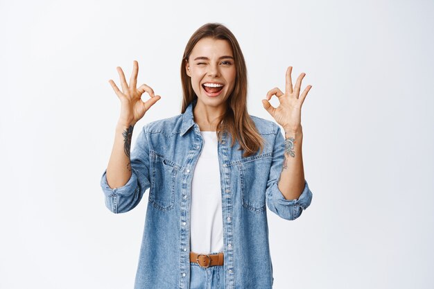 Fille excitée souriante montrant des signes d'accord et un clin d'œil, dites oui ou d'accord, louez un excellent travail, beau geste de travail, debout heureux contre un mur blanc