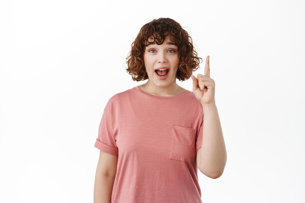 Une fille excitée et surprise crie d'étonnement, pointant le doigt vers le haut, dit wow, vérifiant la publicité sur le dessus, debout en t-shirt sur fond blanc