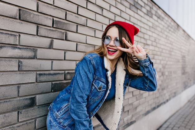 Fille Extatique En Tenue De Denim Drôle Posant Près Du Mur De Briques Au Printemps. Portrait De Joyeux Modèle Féminin Caucasien Debout Dans La Rue Avec Signe De Paix.