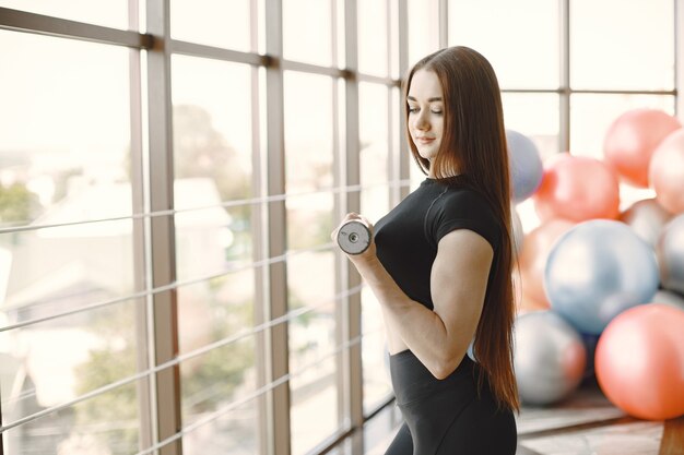 Fille faisant des biceps avec des haltères. Modèle aux cheveux roux en tenue de sport noire dans une salle de sport lumineuse. Balles de fitness en plus.