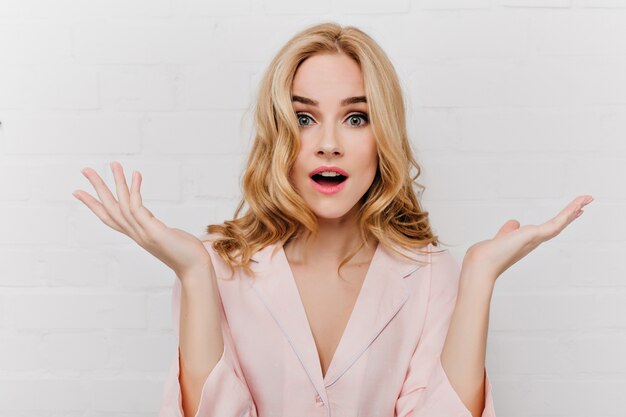 Fille fascinante aux grands yeux bleus exprimant la stupéfaction et agitant les mains isolés sur le mur blanc. Belle femme avec une coiffure frisée porte un pyjama rose posant avec un visage surpris.