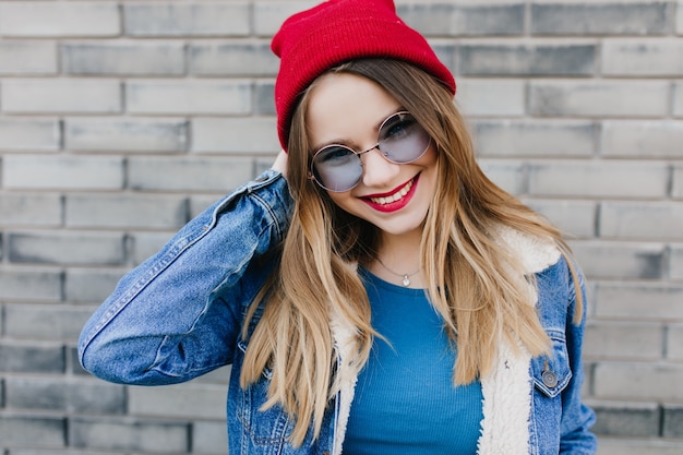 Fille fascinante avec une expression de visage heureux posant dans la rue le matin. Photo extérieure d'une belle dame européenne porte un chapeau rouge souriant