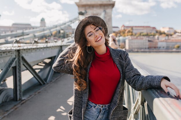 Fille fascinante en pull rouge tricoté souriant joyeusement sur le pont en journée ensoleillée