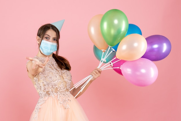 fille de fête avec chapeau de fête et masque médical tenant des ballons colorés sur rose