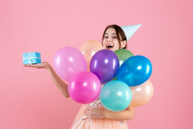 fille de fête avec chapeau de fête tenant des ballons colorés et cadeau sur rose