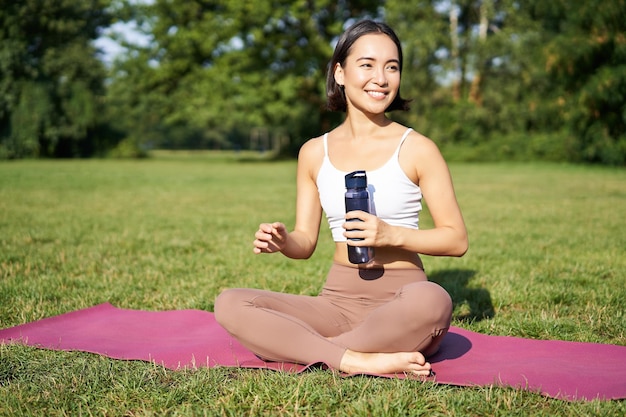 Une fille de fitness souriante est assise sur un tapis et boit de l'eau après l'entraînement de yoga terminé dans les exercices du parc