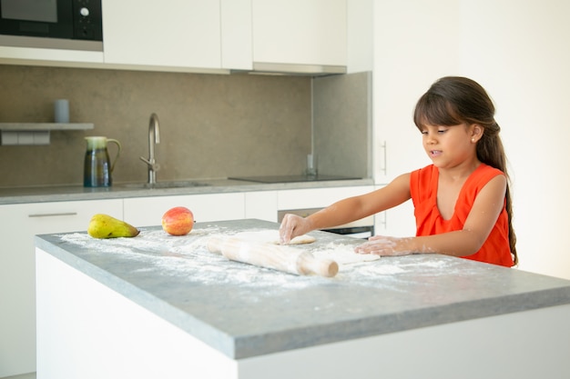 Fille Focalisée Pétrir La Pâte à La Table De La Cuisine. Enfant Faisant Du Pain Ou Un Gâteau Par Lui-même Coup Moyen. Concept De Cuisine Familiale