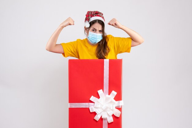 Fille forte avec Bonnet de Noel montrant ses muscles debout derrière un grand cadeau de Noël sur blanc