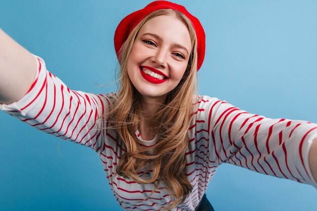 Fille française de bonne humeur prenant selfie avec le sourire. Heureux modèle féminin blonde posant sur le mur bleu.