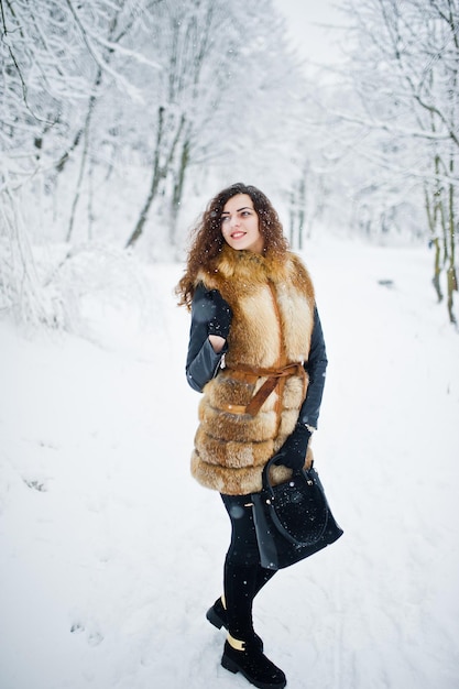 Fille frisée élégante en manteau de fourrure et sac à main au parc forestier enneigé en hiver