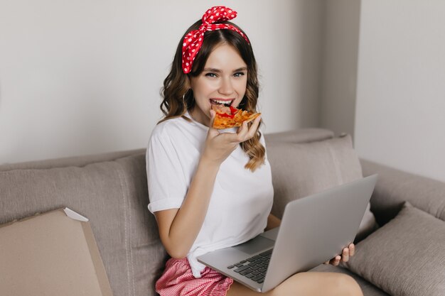 Fille frisée glamour, manger de la pizza avec plaisir et à l'aide d'un ordinateur. Jolie femme passant le week-end à la maison avec ordinateur portable et restauration rapide.