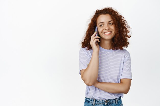 Fille frisée rousse élégante souriante, parlant au téléphone portable avec un visage détendu et insouciant, portant un t-shirt décontracté sur blanc.