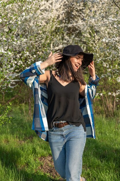Fille gaie dans un chapeau parmi les arbres fleuris au printemps, dans un style décontracté