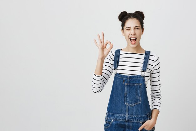 Fille gaie impertinente avec une coiffure de petits pains montrant un geste correct et un clin d'œil, recommander le produit